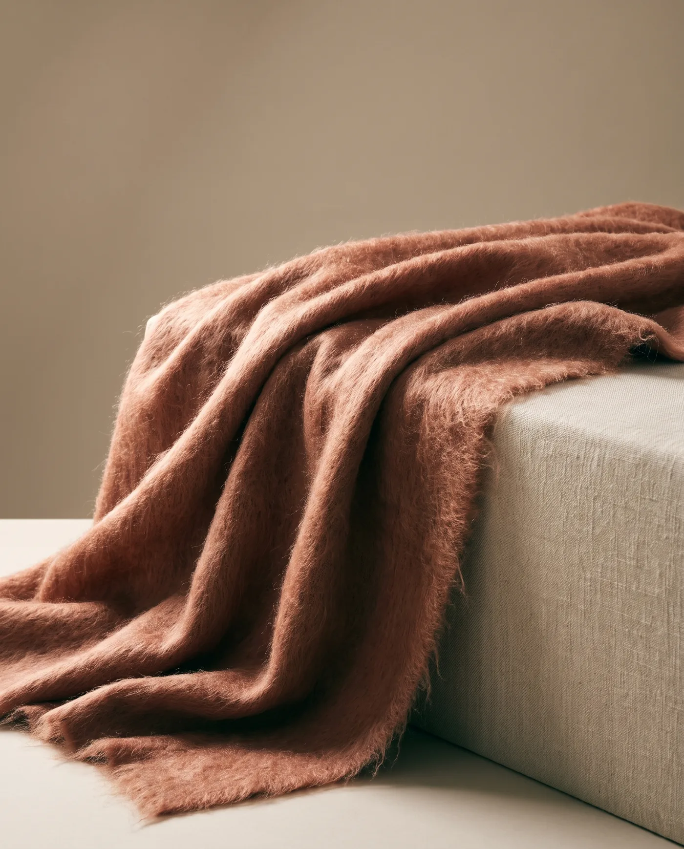 A clay-coloured brushed mohair throw tossed over the corner of a linen plinth, fibres lit from behind.