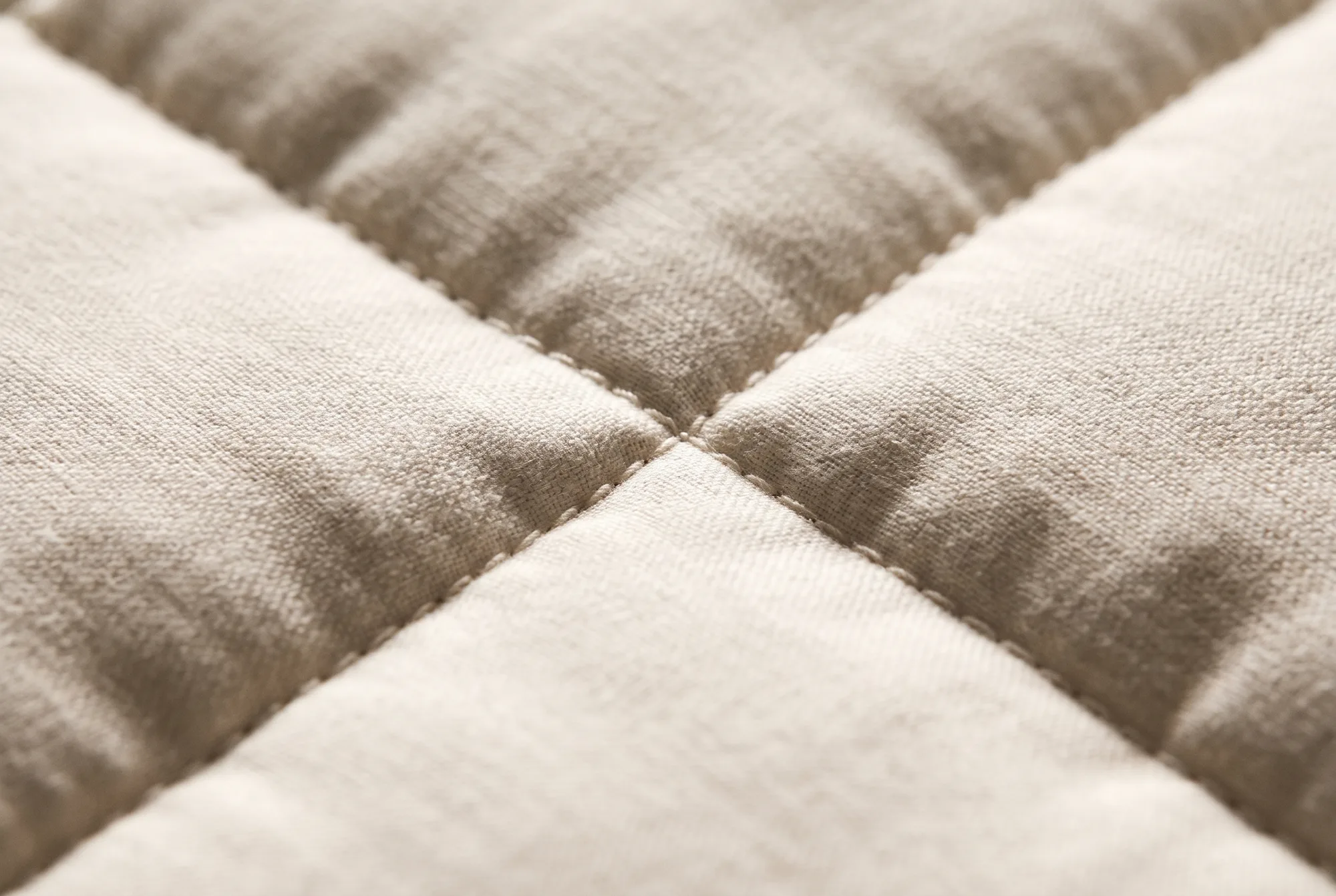 A stitched seam crossing between four quilted squares of bone cotton, glass beads pressing against the weave from inside.